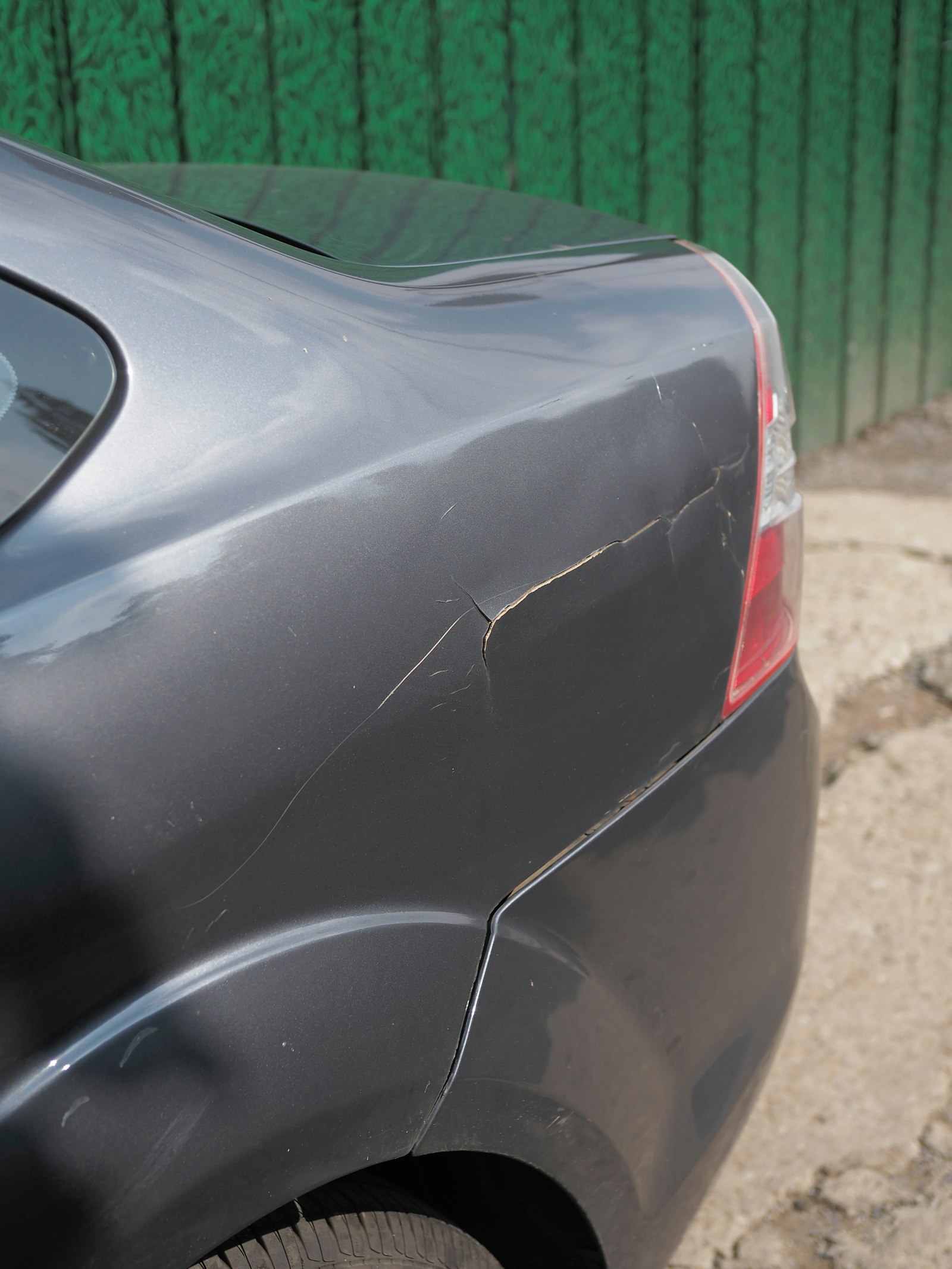 Cracked and flaking paint at the corner of a grey car's rear quarter panel where the bumper seam has split