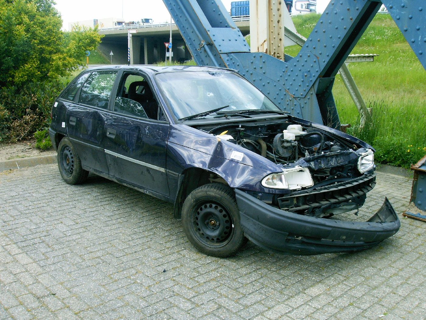 A car with a severely damaged front end, bumper detached, and fender bent past reshaping after a collision