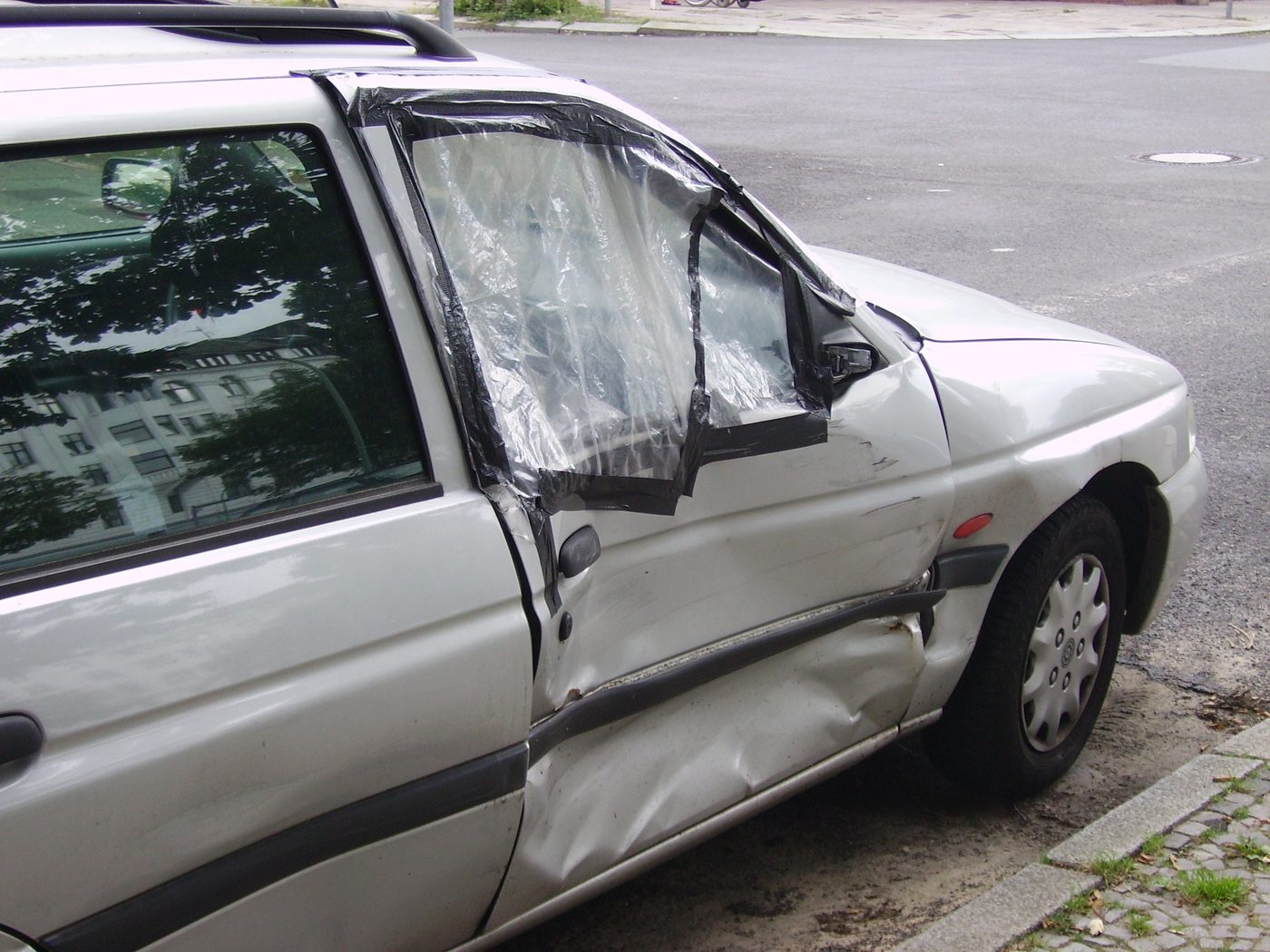 A car door with significant crease damage and a broken window covered in tape after a collision