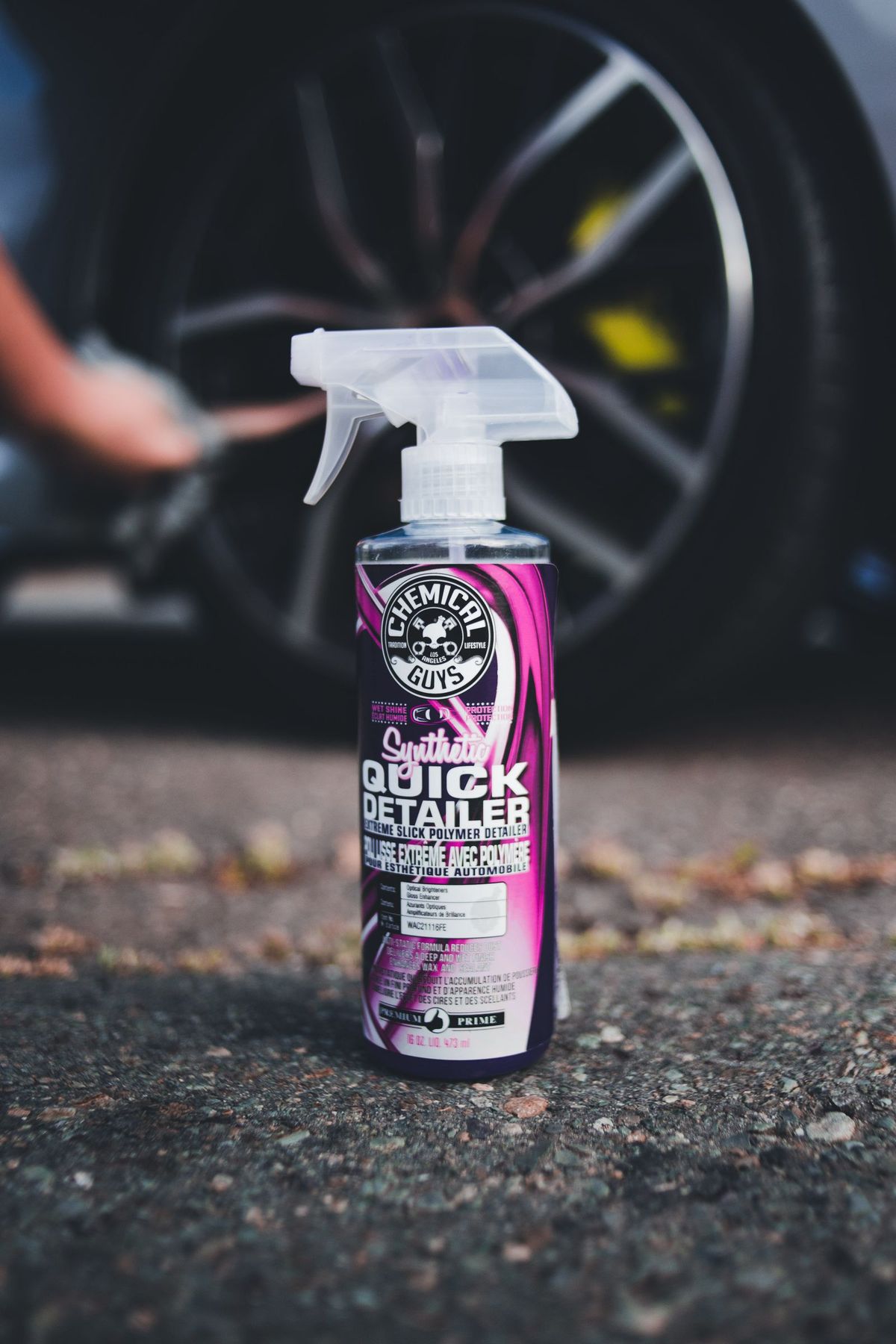 A spray-on automotive product bottle in the foreground with a car wheel visible blurred in the background