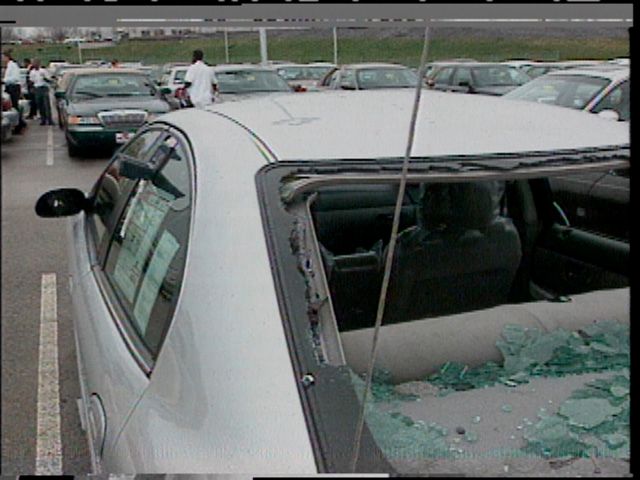 A car in a lot with its side and rear windows shattered by hail, green glass scattered across the seats