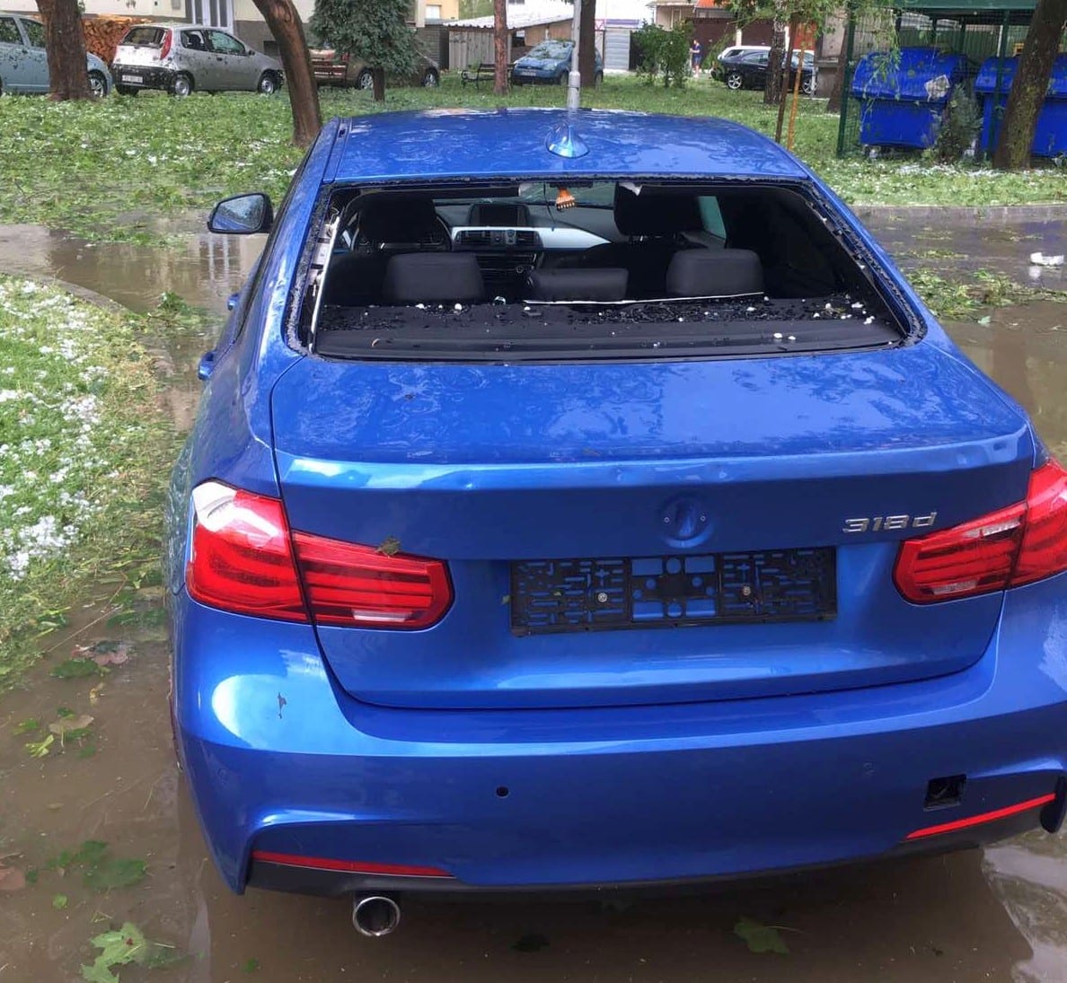 A blue sedan with a dented rear deck and a shattered rear window, hailstones still lying on the grass around it