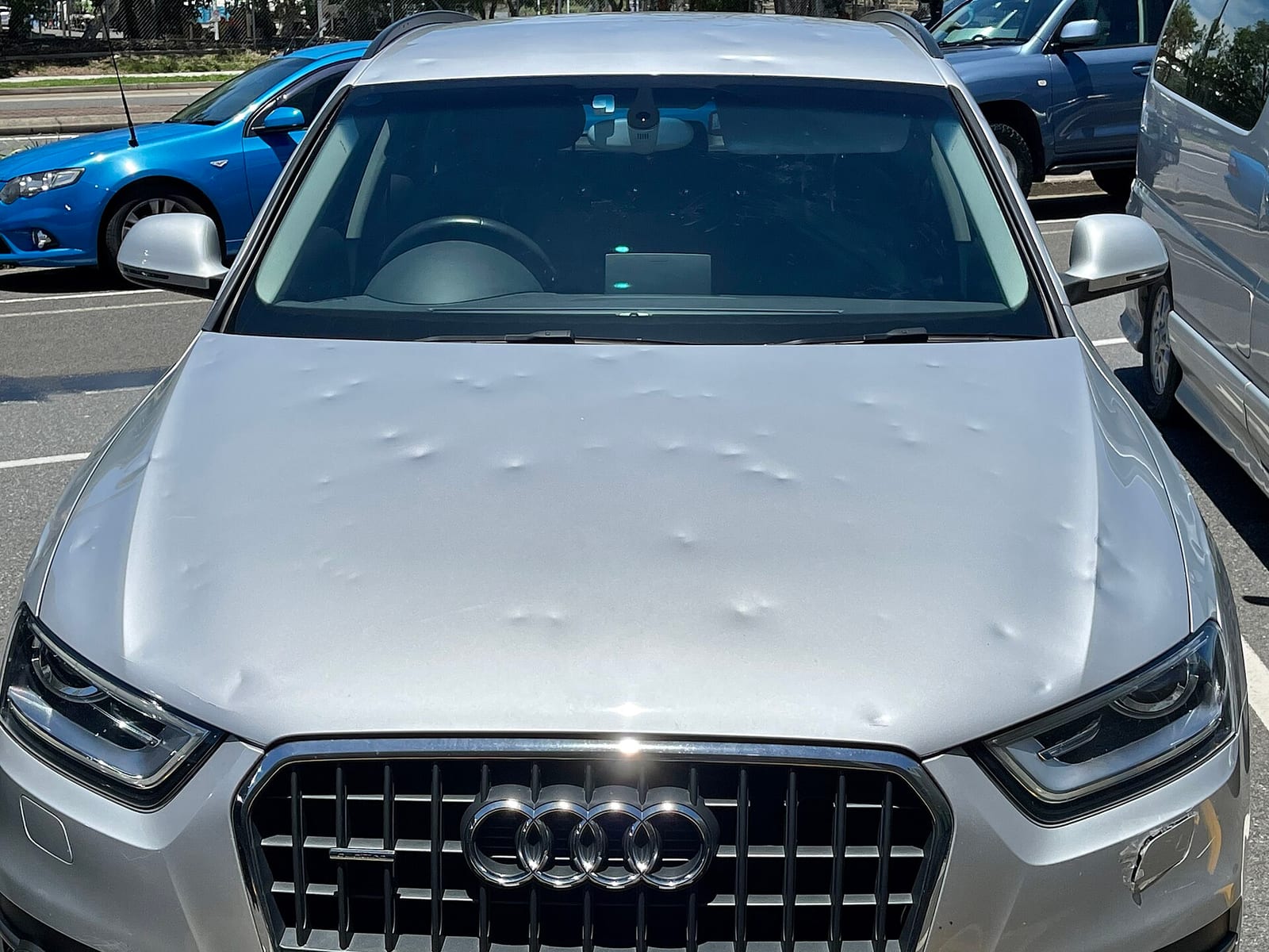 A silver Audi Q3 crossover with dozens of round, paint-intact hail dents across its hood and roof