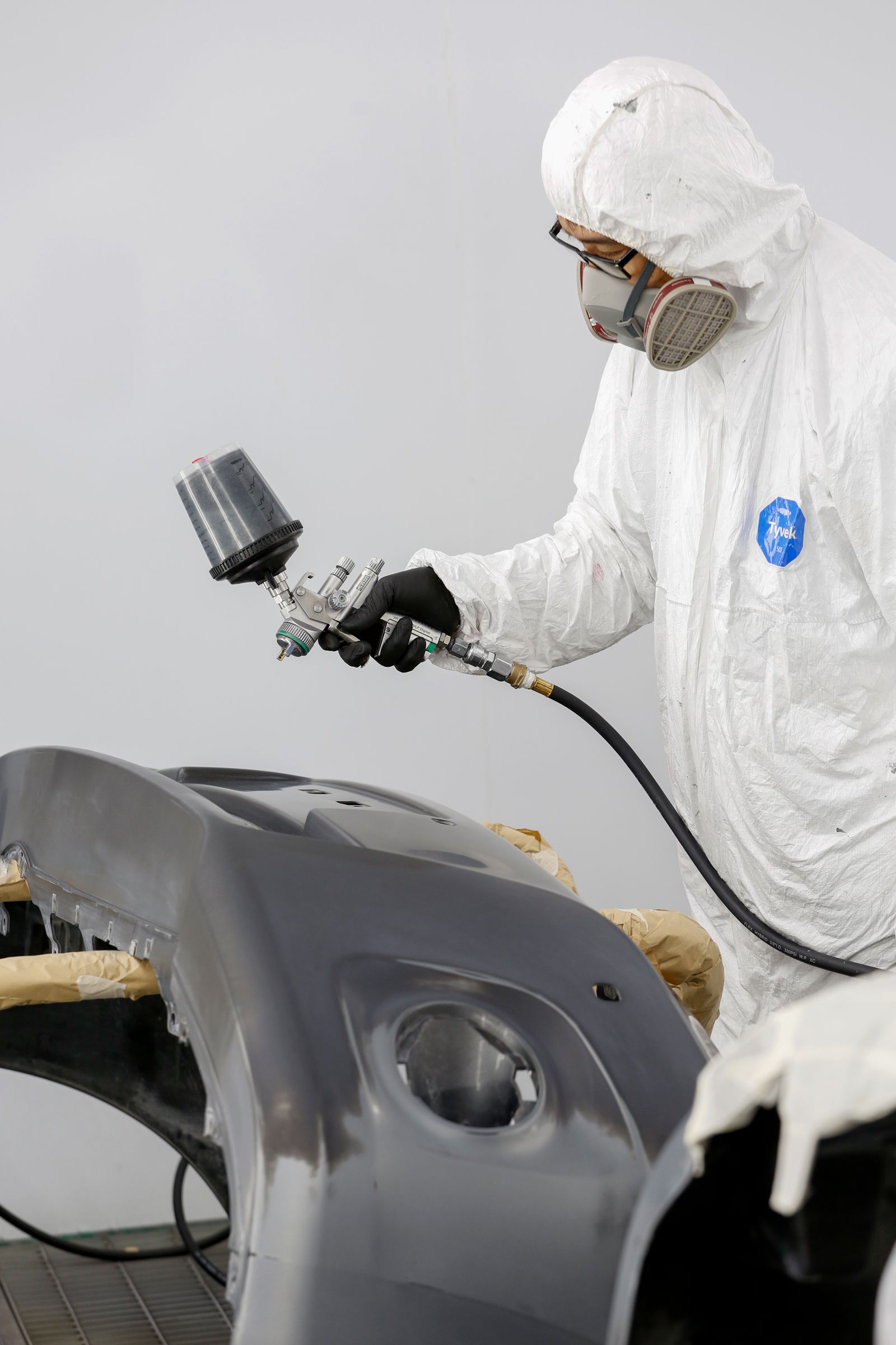 Automotive technician in protective gear spray painting a car bumper in a paint booth