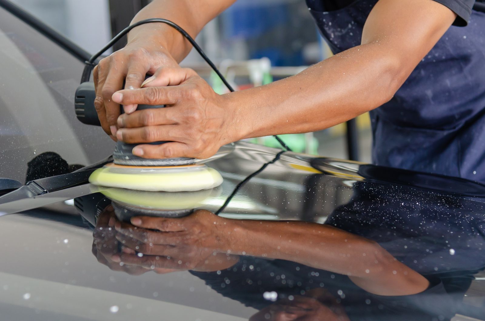 A worker's hands buffing a car hood with an orbital polisher in an auto detailing shop