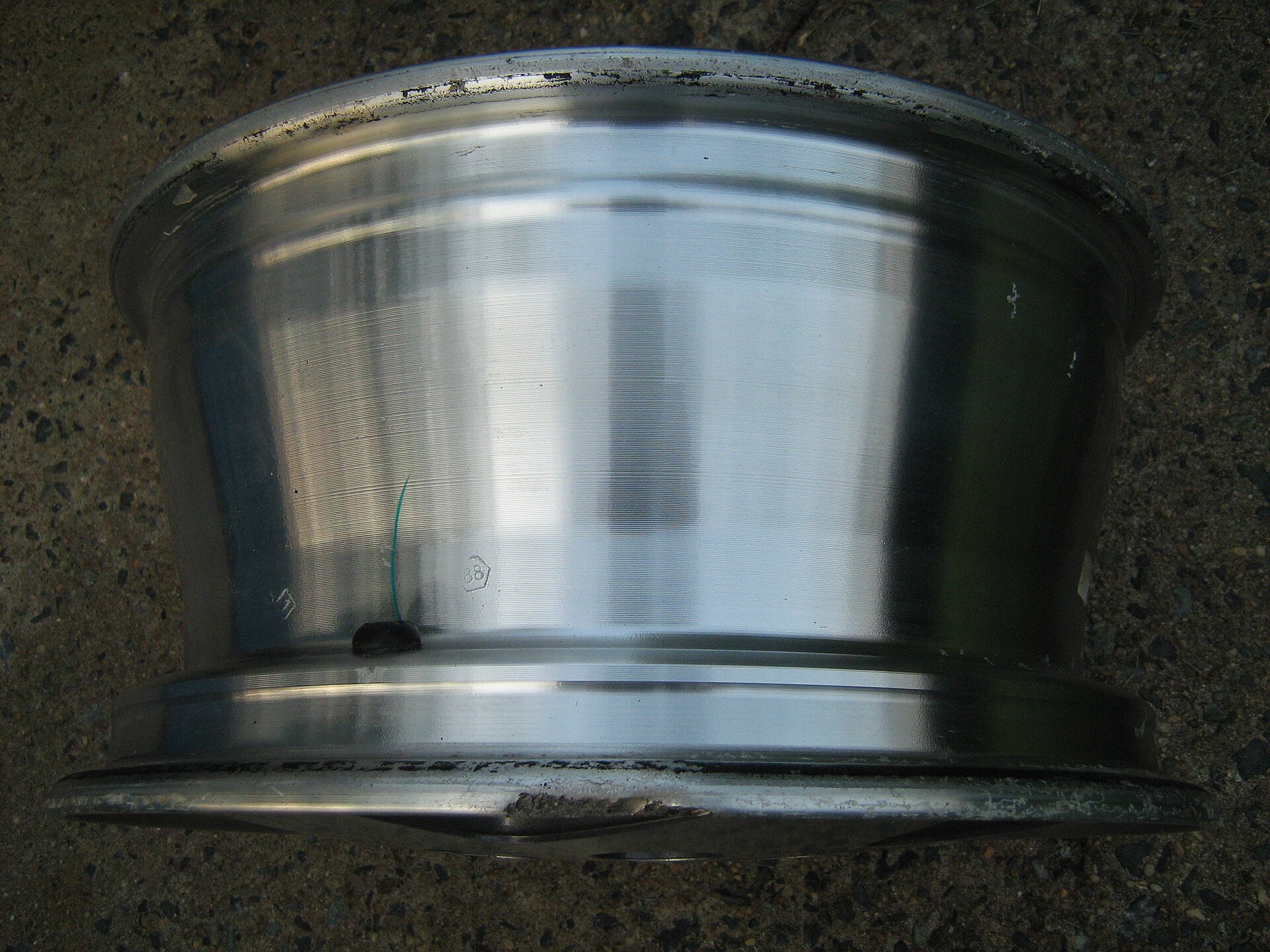 A bare polished alloy wheel lying on gravel with chipped and scuffed dark marks along both outer lip edges